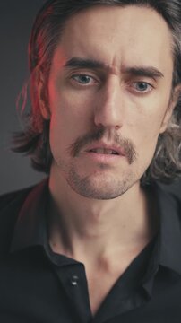 Understanding, realizing, accepting young attractive man with mustache and long hair in a black shirt, dramatically lit by red light against a dark background