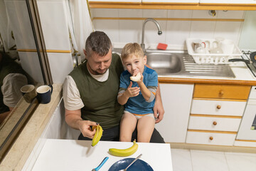 Father feeding son banana in small kitchen