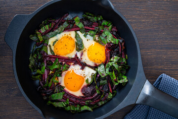 Fried beet stems and eggs in a frying pan on a wooden table, closeup, top view. Food background. Cooked roasted beetroot leaves, eggs with spices for breakfast. Healthy food concept