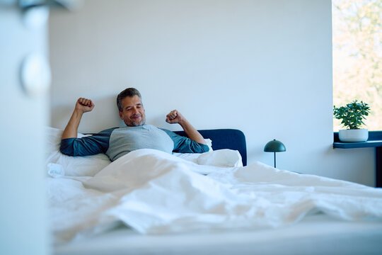Happy man stretching in bed after sleeping, feeling rested and invigorated in his modern bedroom during the morning