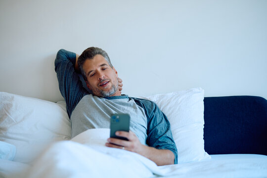 Man lying comfortably in bed, using his mobile phone, browsing applications, connected to technology in the bedroom