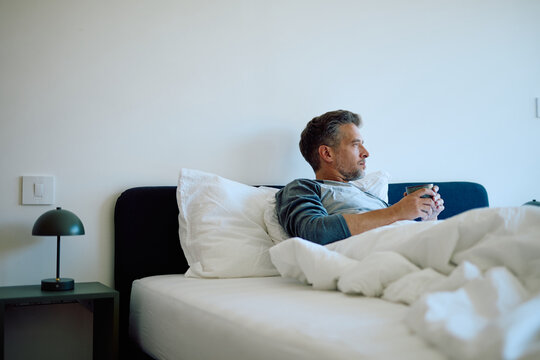Elderly man with grey hair is sitting in bed, clutching a coffee mug and looking lost in thought as he begins his morning routine in a peaceful and comfortable bedroom