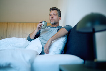 Thoughtful man sitting up in bed, looking away while drinking a hot beverage, enjoying quiet morning leisure time