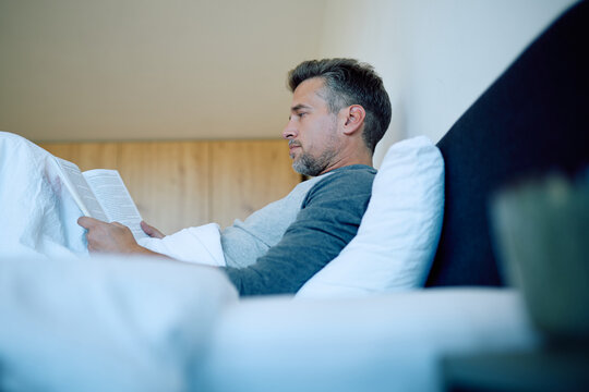 Middle-aged man unwinding in bed in the evening, savoring a peaceful moment with a paperback novel as he prepares for sleep - Powered by Adobe