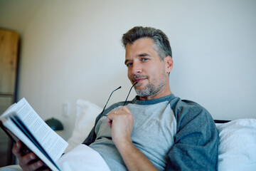 Older gentleman is unwinding in bed, gently holding his reading glasses and a book as he savors a peaceful morning or leisurely weekend activity