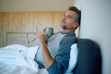 Mature man relaxing in bed, drinking morning coffee and holding glasses, contemplating or thinking deeply