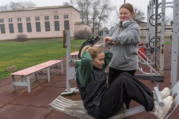 Two young women exercise together on an ab bench at an outdoor gym in a public park, performing crunches while one friend times and supports the other's core training