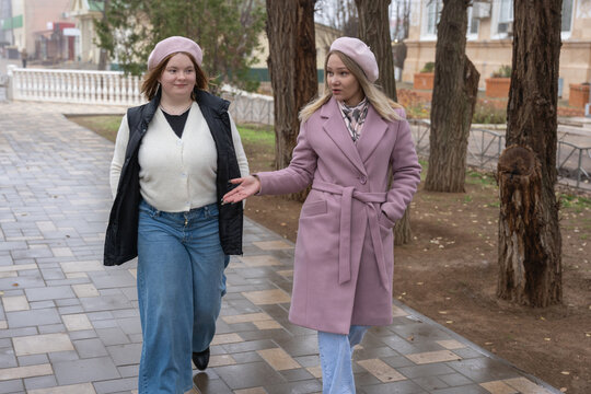 Two women in berets and coats stroll down a fall sidewalk gesturing and arguing during a candid city conversation