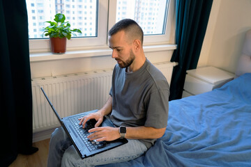 A man is lying down on his bed in a bedroom, focused on typing on his laptop. He appears to be working remotely while taking a break from his regular routine