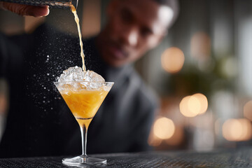 A bartender pours a vibrant orange cocktail over ice into a martini glass. The drink evokes sophistication and refreshment. Ideal for food, beverage, and lifestyle content.