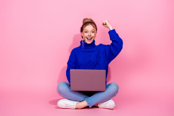 Young woman with blue sweater sits on a pink background using laptop smiling excitedly celebrating...
