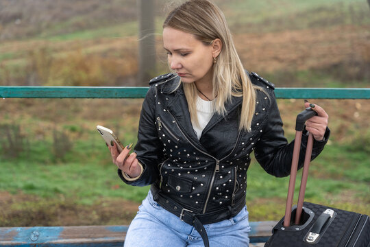 Solo traveler sitting on bench at rural bus stop with luggage using smartphone