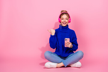 Young woman wearing blue knit sweater sits with pink headphones and phone against pink background...