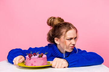 Young woman in blue sweater sits at a table with a pink cake on a pink background looking annoyed