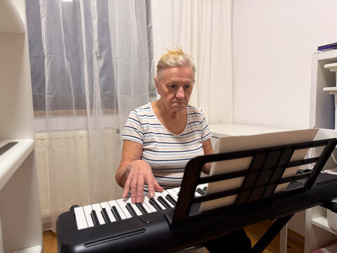 An elderly woman playing a keyboard in a bright room, focused on her music sheet - Powered by Adobe