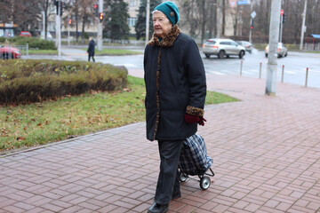 Fototapeta premium Elderly woman on street. Pensioner walks with cart for things. Woman walks on road. Transportation of personal belongings.