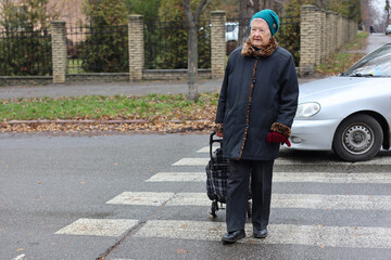 Fototapeta premium Elderly woman on street. Pensioner walks with cart for things. Woman walks on road. Transportation of personal belongings.
