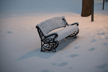 Snow-Covered Park Bench in Quiet Winter Scene with Footprints and Soft Evening Light