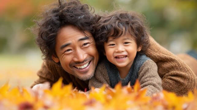 On a bright, sunny afternoon, a father and his young son joyfully lie in a blanket of vibrant autumn leaves. Their laughter fills the air as they share a tender moment together