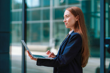 Business professional stands confidently with laptop outside modern office building in urban setting © Iryna