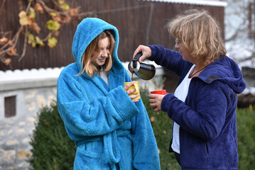Mother pours coffee from coffee percolator for her young adult daughter dressed in blue bathrobe in the garden