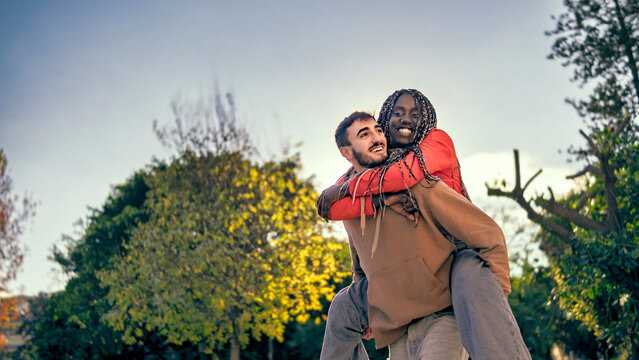 Joyful interracial couple sharing playful piggyback moment outdoors at sunset. Authentic expressions, modern lifestyle vibes. Love, happiness, diversity, togetherness, youth and positive relationships