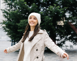 Young woman with long dark hair, wearing a cozy white beanie and beige trench coat, joyfully...