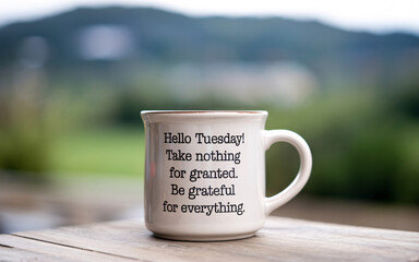 A white mug with a tuesday message sitting on a wooden surface with a blurred green background
