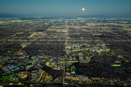 Aerial view of illuminated Los Angeles grid at night under a full moon, showing vast urban sprawl, streets, and city lights extending to the horizon across Southern California