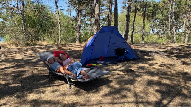 A mother and son relax on a lounger near a blue tent. They lie together in a sunny pine forest surrounded by nature during a camping trip.