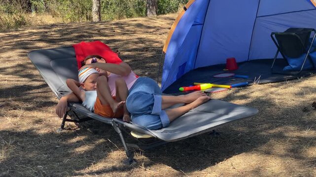 A mother and son relax on a lounger near a blue tent. They lie together in a sunny pine forest surrounded by nature during a camping trip.