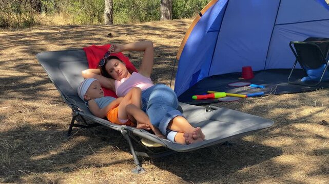 A mother and son relax on a lounger near a blue tent. They lie together in a sunny pine forest surrounded by nature during a camping trip.