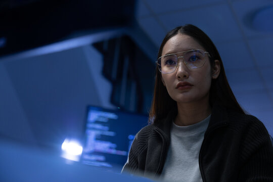 Young woman wearing glasses focused at computer screen in dim office with code visible on monitor - Powered by Adobe