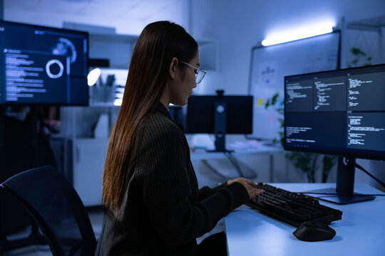 Young woman programmer coding at computer in dark office with multiple monitors and blue light, focused and concentrated - Powered by Adobe