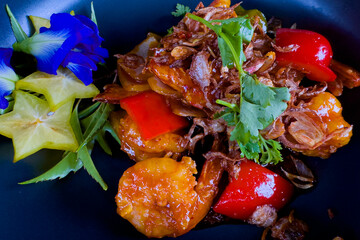 Tamarind sauce shrimp topped with crispy garlic and red bell peppers, served on a black plate, with starfruit and butterfly pea flowers arranged on the side.