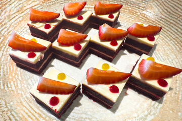 These are small layered dessert squares with a dark cake base and a pale creamy topping. Each piece is decorated with a slice of strawberry and colorful syrup drops.