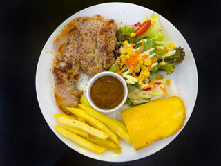 A plate holds a grilled pork chop seasoned with pepper, accompanied by a side salad, golden French fries, and a slice of toasted garlic bread.