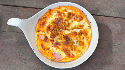 he pizza is cut into slices and features a golden-brown, melted cheese top with visible bits of ham and possibly pineapple. It is presented on a rustic wooden table.