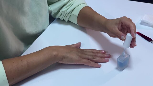 A woman applies clear polish to her fingernails using a brush at a white table. She carefully paints her nails during a manicure session with beauty tools nearby.
