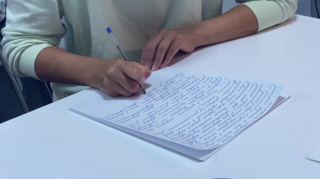 A woman writes a long text on a sheet of paper using a blue pen. She sits at a white table while focusing on her handwriting during a study session.