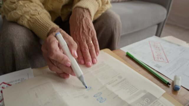 Tilt up of senior woman highlighting overdue household bills while reviewing expenses and managing personal finances on wooden table at home in realistic domestic setting