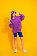 Young woman in purple hoodie holding tennis racket smiling against a bright yellow background