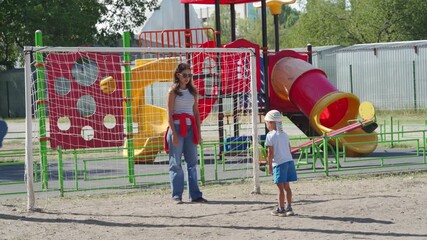 A happy mother plays goalkeeper while her son kicks a soccer ball. They enjoy a fun game at the outdoor playground on a sunny summer day. - Powered by Adobe