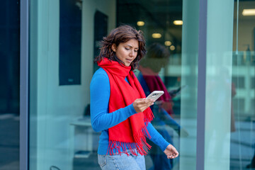 Woman in red scarf checks her phone while walking near glass building during daytime