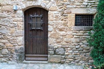Rustic wooden arched door set within an aged stone wall in the pedestrian historic, fortified medieval town Peratallada, Girona, Spain. Classic old facade texture.
