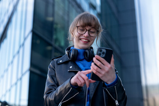Young woman with headphones smiles while using smartphone outside modern building in city - Powered by Adobe
