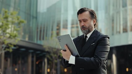 Portrait of old businessman scrolls newsfeed on digital tablet in downtown. Entrepreneur with grey beard checks market trends on internet via mobile device against business center - Powered by Adobe