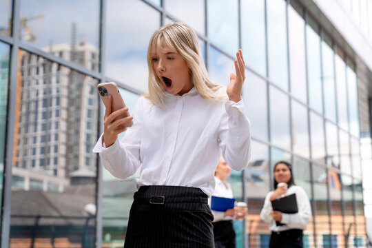 Woman in business attire reacts with surprise while checking her phone outside a modern office building