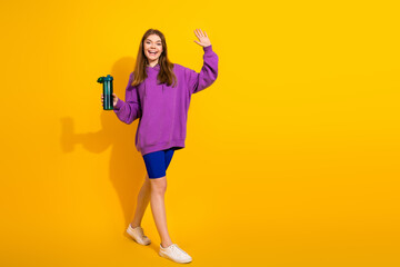 Young woman in purple hoodie waves and smiles holding a water bottle against a bright yellow background