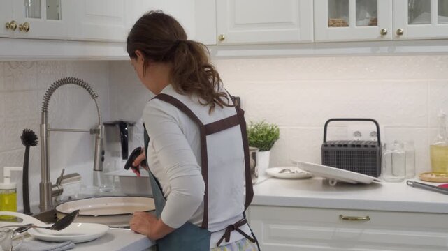A busy housewife washes a large enamel pot at the sink. She cleans dirty cookware after cooking in her bright modern kitchen.